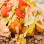 A close-up of a Big Mac Tater Tot Casserole with ground beef, tater tots, shredded lettuce, diced tomatoes, chopped pickles, and creamy sauce, served on a white plate.