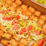 The baked in a rectangular pan, topped with rows of crispy tater tots, shredded lettuce, chopped tomatoes, sliced pickles, and sesame seeds. Small glass bowls of sesame seeds and pickles are nearby.