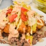 A plate of Big Mac Tater Tot Casserole features ground beef, onions, tater tots, shredded lettuce, tomato, pickles, and creamy sauce, with bowls of pickles and sesame seeds in the background.