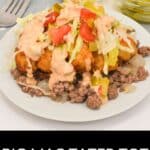 A plate of Big Mac Tater Tot Casserole is topped with chopped lettuce, tomatoes, pickles, and creamy sauce, with bowls of sesame seeds and extra pickles in the background.