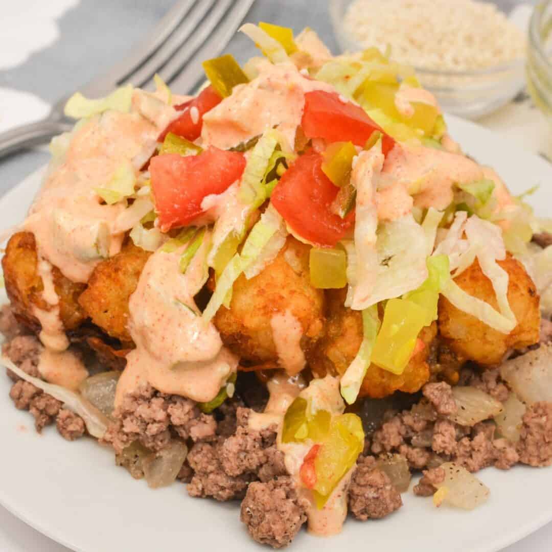 A plate of seasoned ground beef and onions, inspired by Big Mac Tater Tot Casserole, topped with tater tots, shredded lettuce, diced tomatoes, sliced peppers, and creamy sauce.