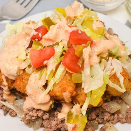 A plate of ground beef topped with fried dough pieces, chopped lettuce, tomatoes, pickled peppers, and a creamy sauce—reminiscent of a Big Mac Tater Tot Casserole—sits next to a fork and a small bowl of sesame seeds.