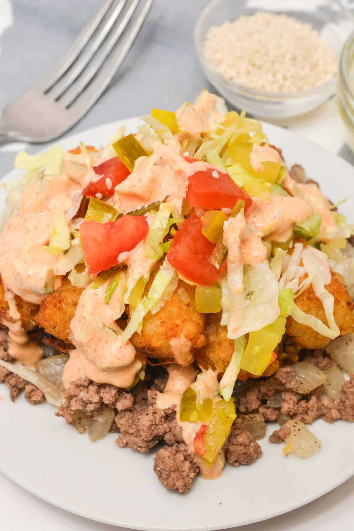 A plate of ground beef topped with fried dough pieces, chopped lettuce, tomatoes, pickled peppers, and a creamy sauce—reminiscent of a Big Mac Tater Tot Casserole—sits next to a fork and a small bowl of sesame seeds.