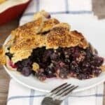 A slice of blueberry pie on a white plate sits on a blue and white checkered napkin. Text reads, "19 Blueberry Recipes Perfect for Summer—refreshing blueberry dishes you'll want to try for your summer desserts.