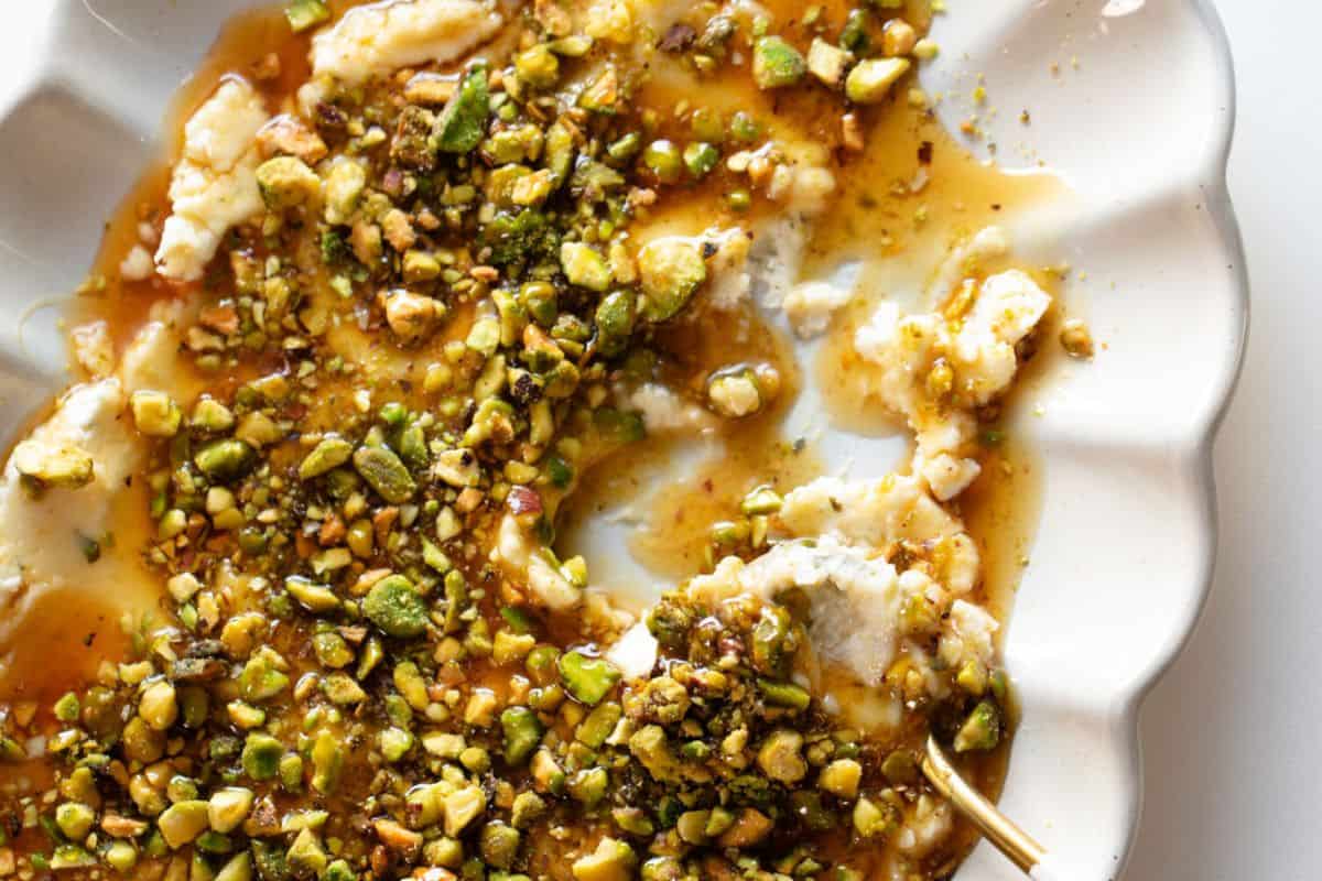 A white plate with creamy cheese topped with chopped pistachios and honey, partially eaten, with a gold fork on the side—an elegant choice for those seeking cold appetizer ideas.