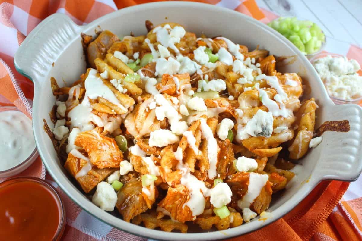 A bowl of loaded fries topped with buffalo chicken, ranch dressing, and crumbled blue cheese, with small bowls of dipping sauces and chopped celery on the side—this is a crazy good addition to your favorite French fry recipes.