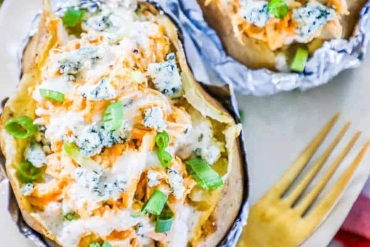 Discover unique baked potatoes with this recipe: baked potatoes topped with shredded chicken, blue cheese crumbles, green onions, and creamy dressing, served on a plate with a gold fork—perfect for new baked potato ideas.