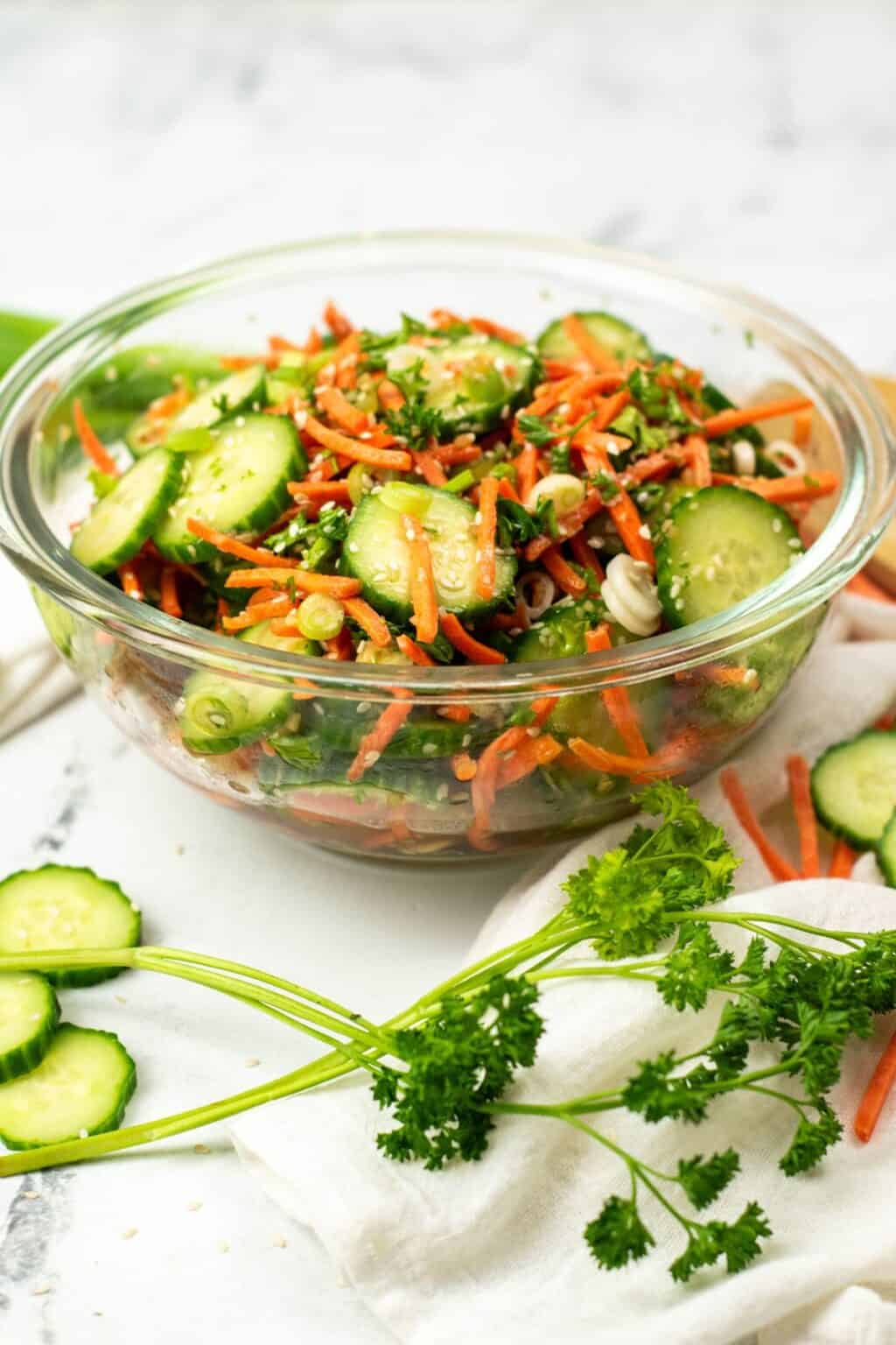 A glass bowl filled with a fresh Carrot and Cucumber Salad—sliced cucumbers, shredded carrots, green onions, and herbs—garnished with cucumber slices and parsley on the side.