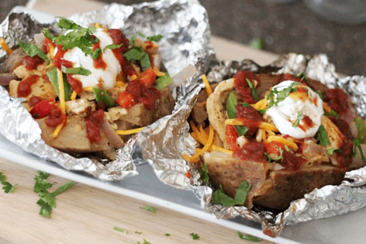 Two baked potatoes in foil topped with shredded cheese, fajita chicken, salsa, sour cream, and chopped cilantro—served on a white plate—make for delicious baked potato ideas to try at home.