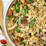 A glass bowl filled with Salad featuring rotini, spinach, tomatoes, cucumber, onions, and shredded cheese, all garnished with fresh herbs.