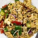 A white plate with Chicken and Pesto Pasta Salad featuring rotini, cherry tomatoes, spinach, red onions, pine nuts, mozzarella, chicken, and cucumber, all topped with shredded cheese.