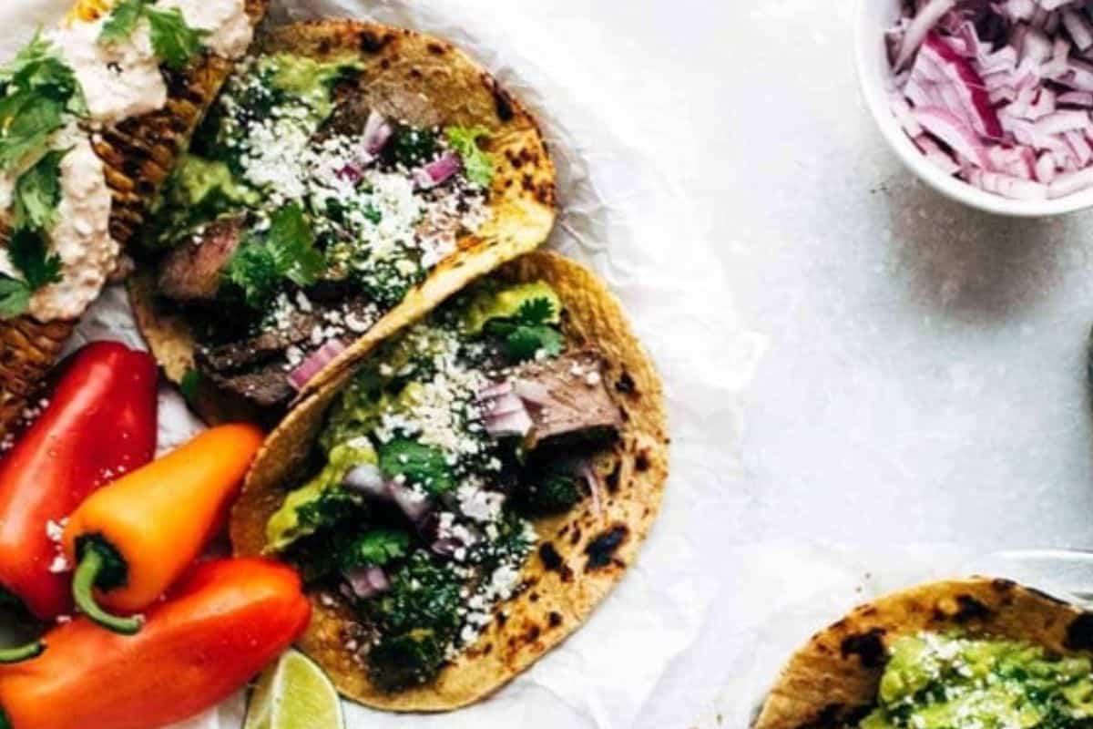 Two tacos with juicy steak, guacamole, onions, cheese, and cilantro on tortillas, served alongside mini bell peppers and a bowl of chopped red onions—a delicious nod to steakhouse favorites.