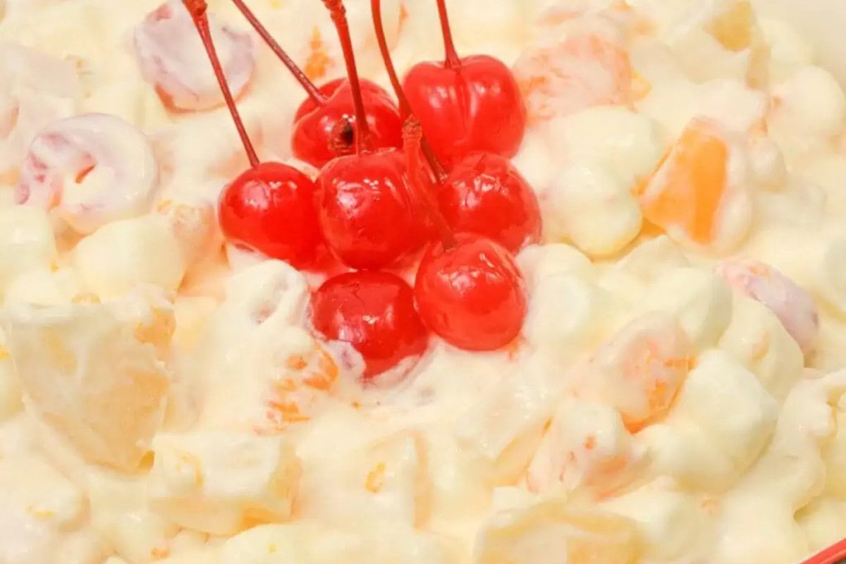 A close-up of ambrosia salad with mini marshmallows, fruit pieces, creamy dressing, and several maraschino cherries on top—one of the most beautiful fruit salads you'll love to try.