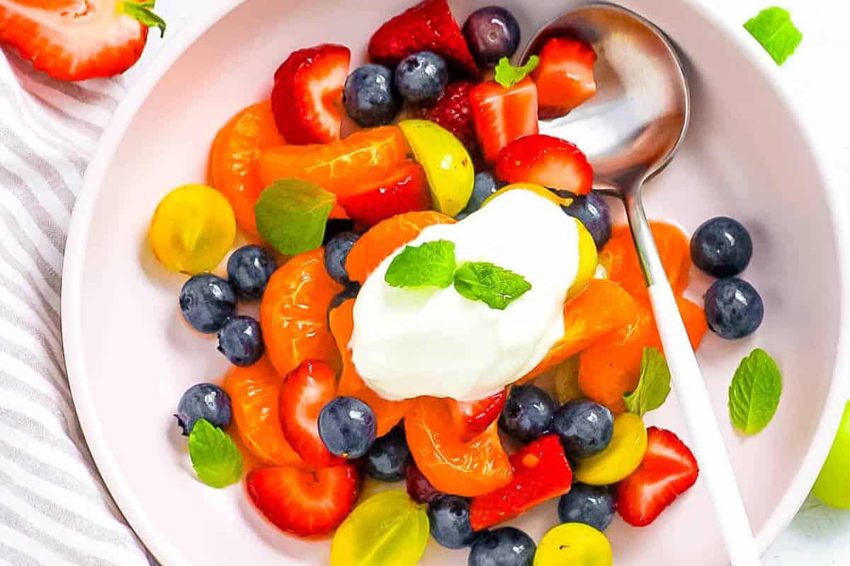 Bowl of fresh fruit salad with strawberries, blueberries, tangerine segments, and yellow tomatoes, topped with yogurt and mint leaves—a perfect example of Beautiful Fruit Salads, served with a spoon on the side.