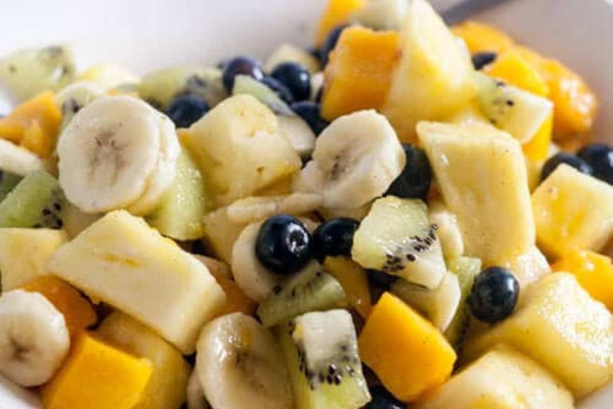 A close-up of a delicious fruit salad containing sliced bananas, kiwi, mango, and whole blueberries in a white bowl.