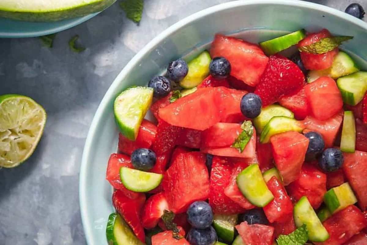 A bowl of beautiful fruit salad featuring watermelon, strawberries, blueberries, cucumber, and mint leaves, with a halved lime nearby.