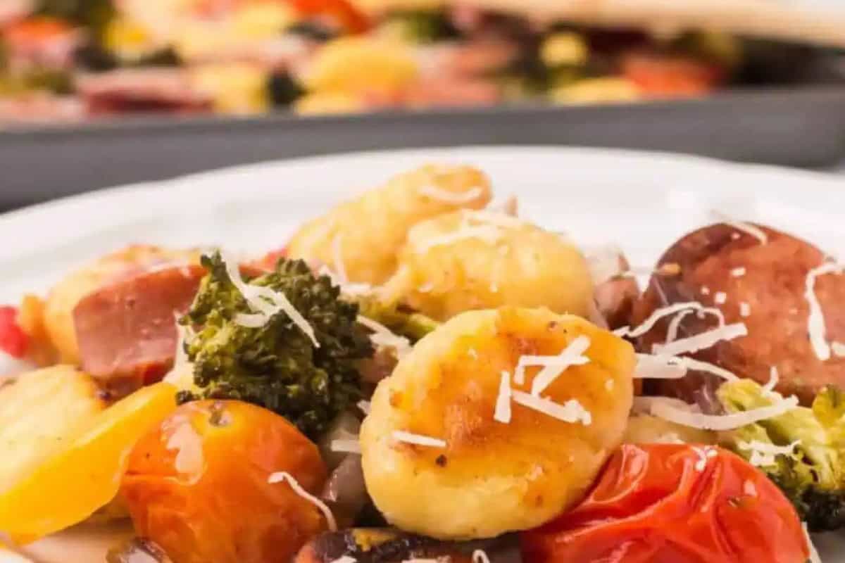 A close-up of a plate featuring gnocchi, broccoli, tomatoes, sausage slices, and grated cheese—an inviting taste of Homemade Italian Recipes—with a similar dish softly blurred in the background.