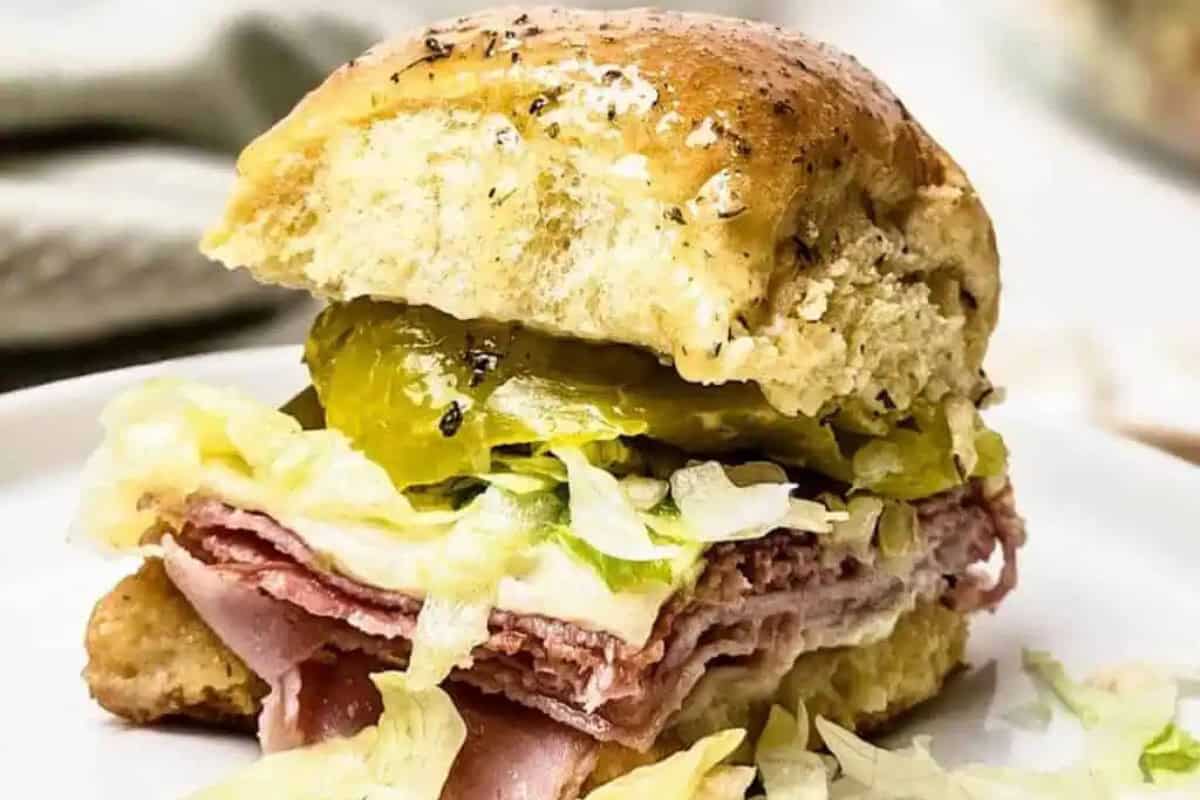 A homemade sandwich on a seeded bun filled with slices of meat, shredded lettuce, pickles, and melted cheese, placed on a white plate—just like Nonna used to make.