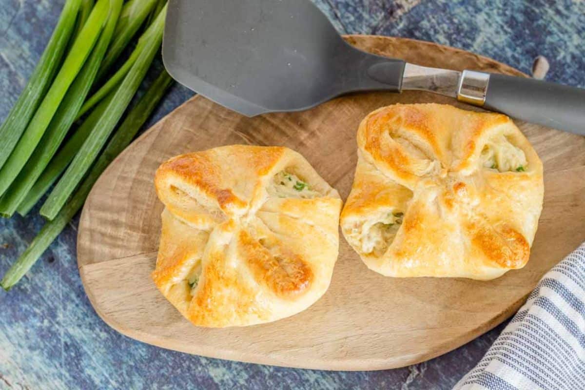 Two baked pastry bundles with a golden crust sit on a wooden board next to a spatula and fresh green onions—perfect for budget friendly dinners using canned chicken.