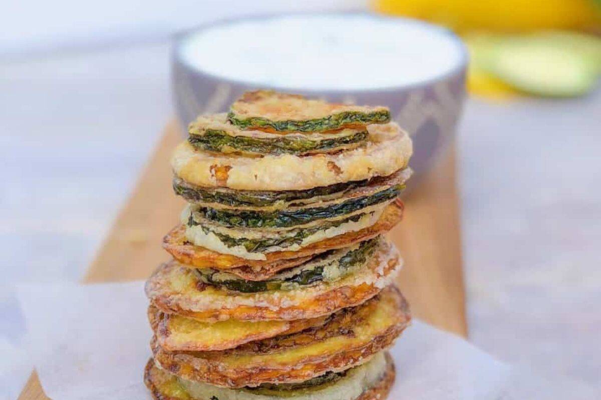 A stack of crispy, breaded zucchini chips sits on parchment paper with a bowl of dip blurred in the background, making them perfect for those seeking unique cold appetizer ideas.