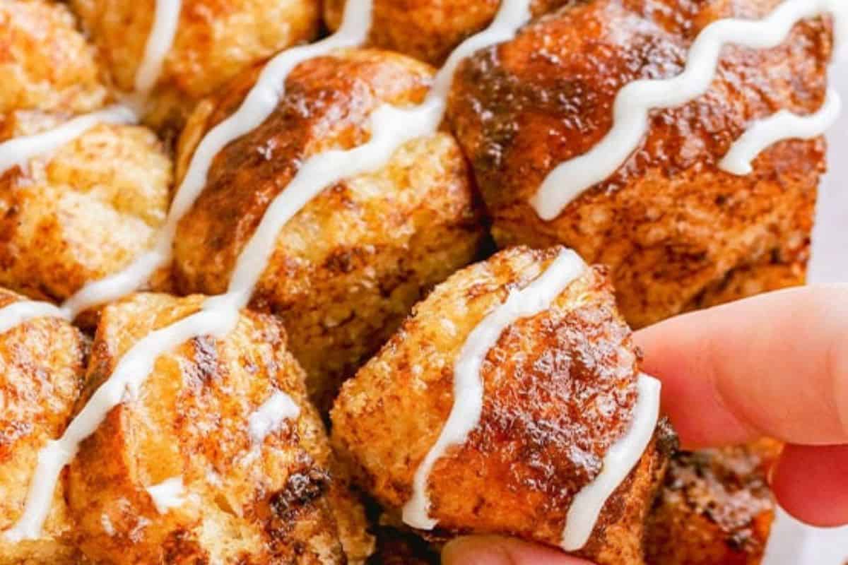 Close-up of baked cinnamon pull-apart bread pieces with a white icing drizzle, one piece being picked up by hand—an irresistible treat perfect for fans of effortless desserts and easy slow cooker desserts.