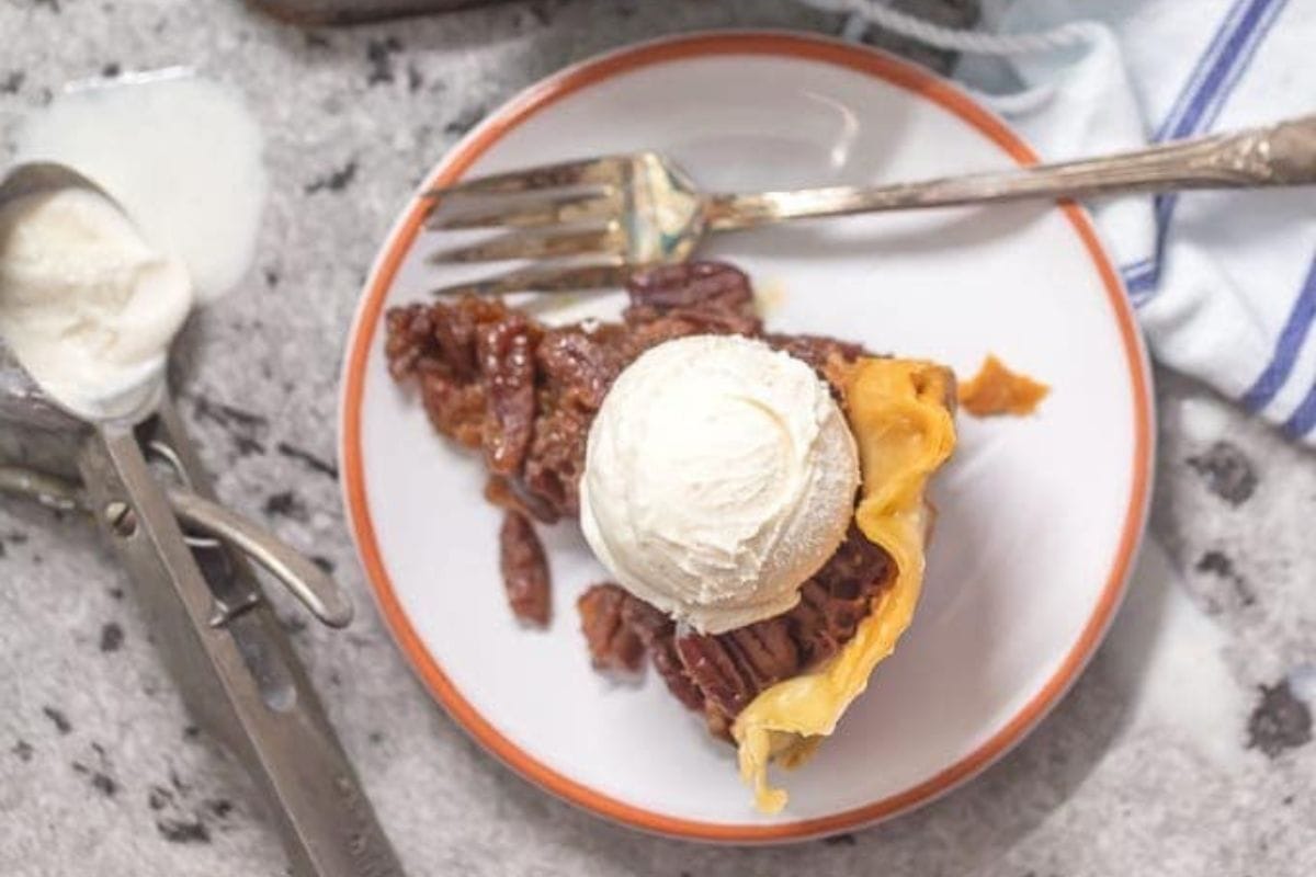 A slice of pecan pie with a scoop of vanilla ice cream on top sits on a white plate with an orange rim, showcasing effortless indulgence—perfect for fans of crock pot desserts or slow cooker desserts.