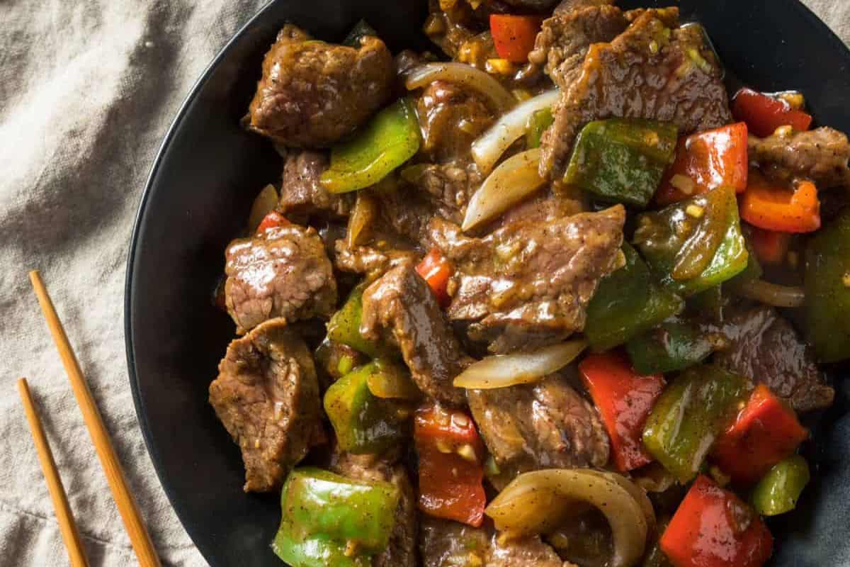A plate of juicy steak stir-fried with green and red bell peppers, onions, and savory sauce sits next to a pair of wooden chopsticks—a delicious twist on classic steak recipes.