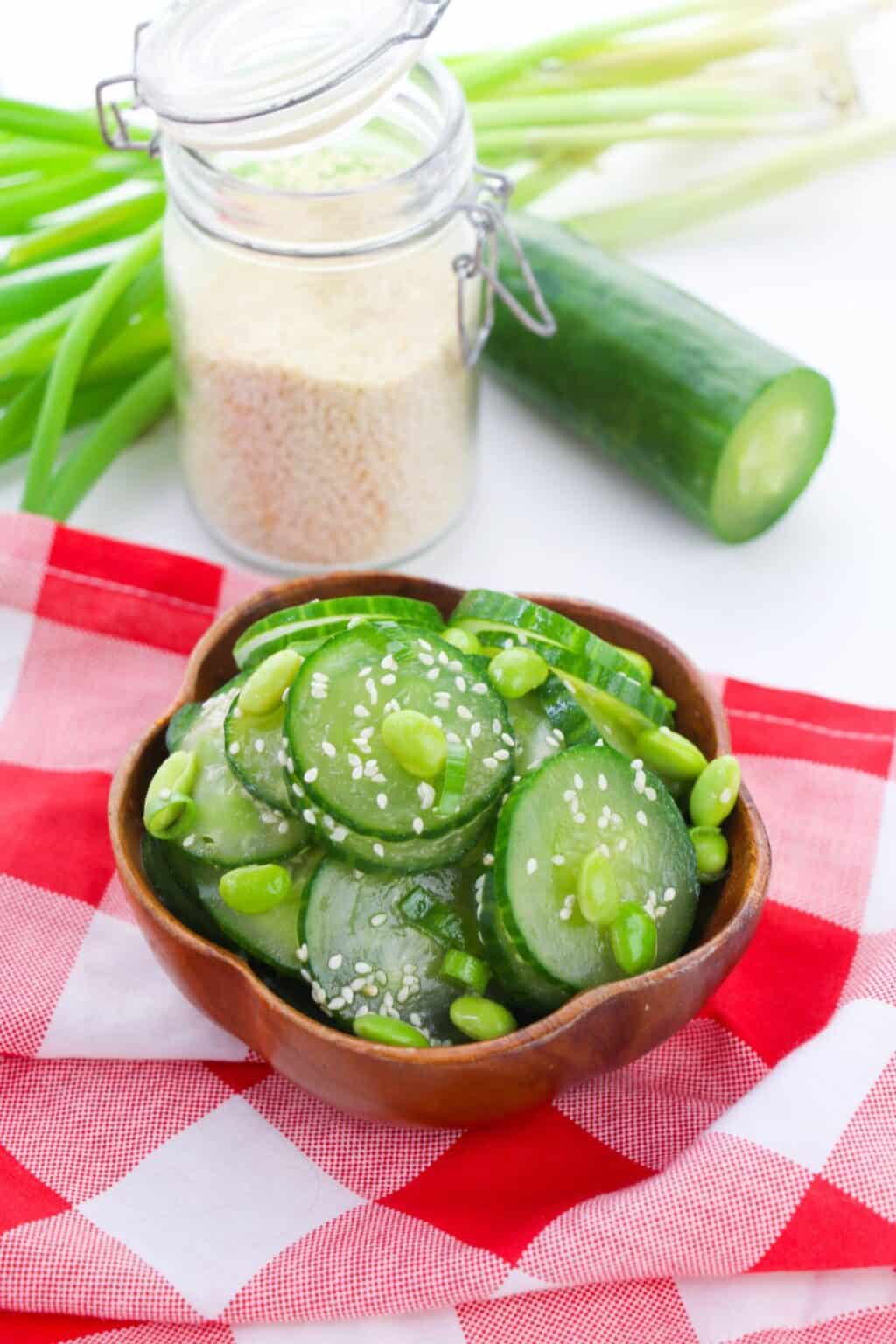A wooden bowl of Cucumber Edamame Salad topped with sesame seeds sits on a red and white checkered cloth, with cucumber, green onions, and a jar of sesame seeds in the background.