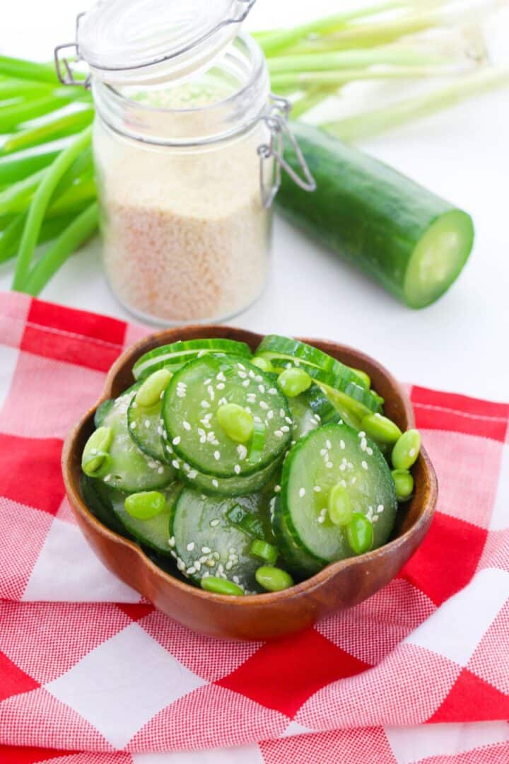 A wooden bowl of Cucumber Edamame Salad topped with sesame seeds sits on a red and white checkered cloth, with cucumber, green onions, and a jar of sesame seeds in the background.