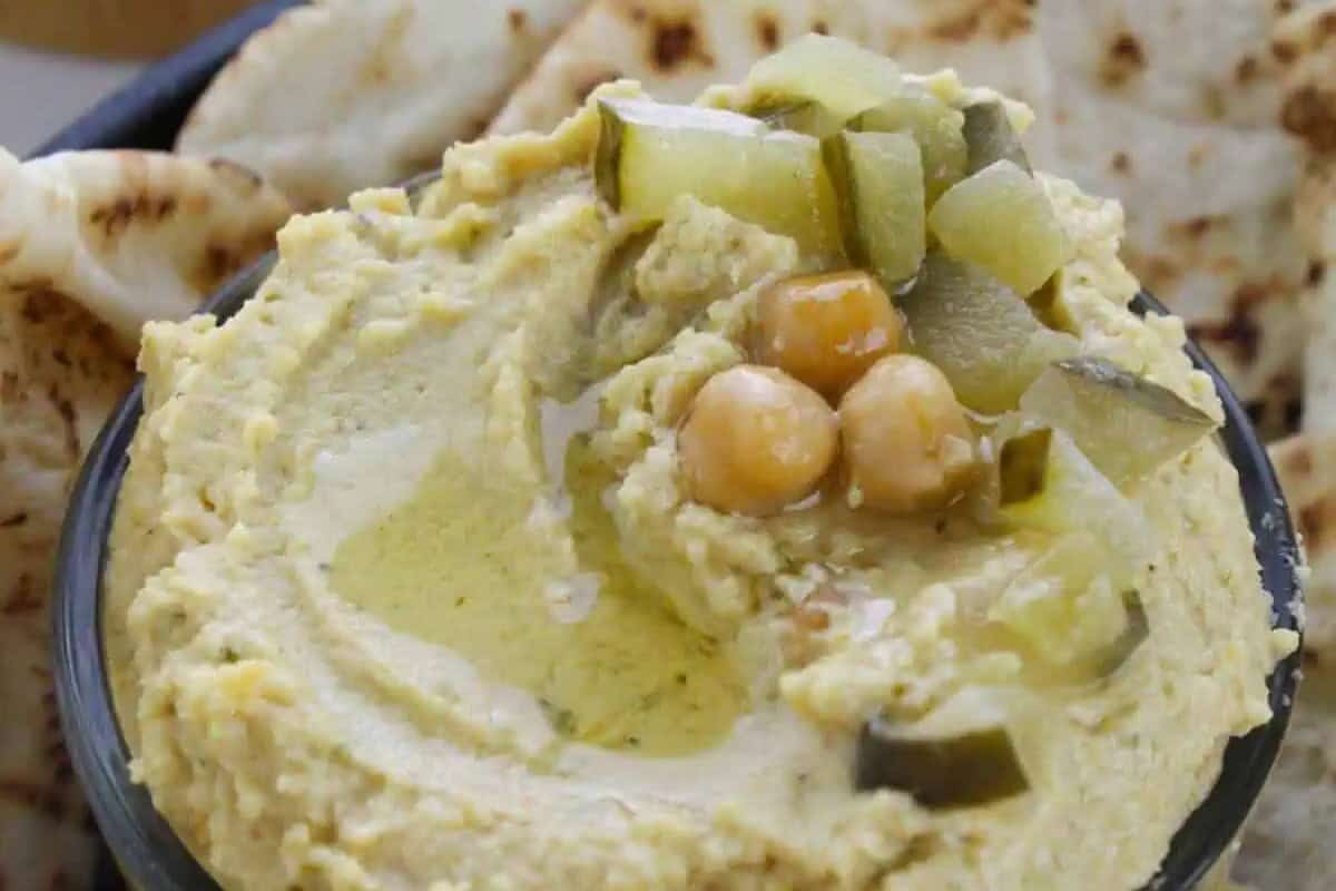A bowl of classic hummus topped with olive oil, chickpeas, and chopped pickles offers delicious twists, served with pita bread in the background.