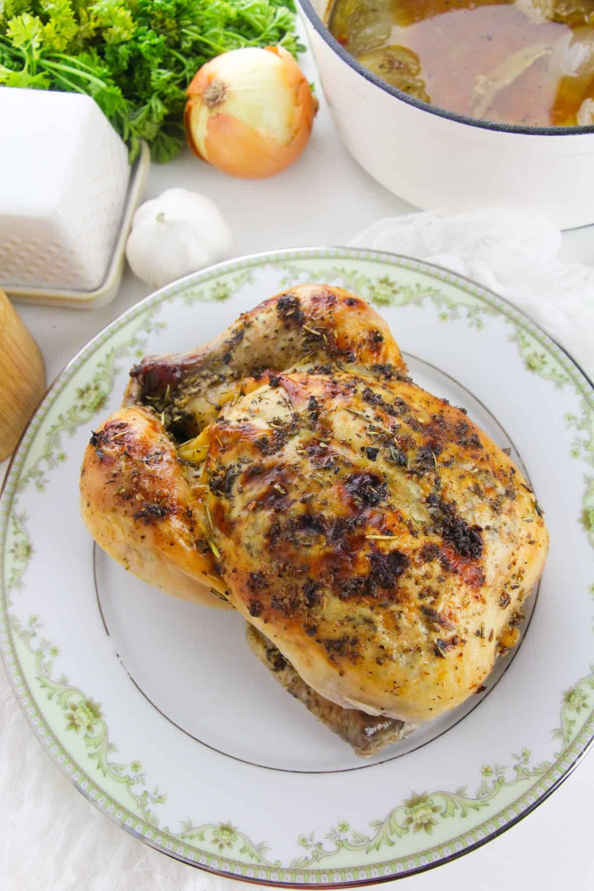 A Dutch Oven Roasted Chicken with herbs sits on a decorative plate; garlic, onion, fresh herbs, and a pot of broth are in the background.