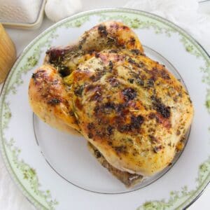 A Dutch Oven Roasted Chicken, seasoned with herbs, is served on a white and green decorative plate.