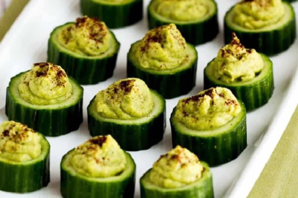 Sliced cucumber rounds topped with a dollop of hummus and a sprinkle of ground spice, arranged neatly on a white rectangular plate—a refreshing addition to your cold appetizer ideas.