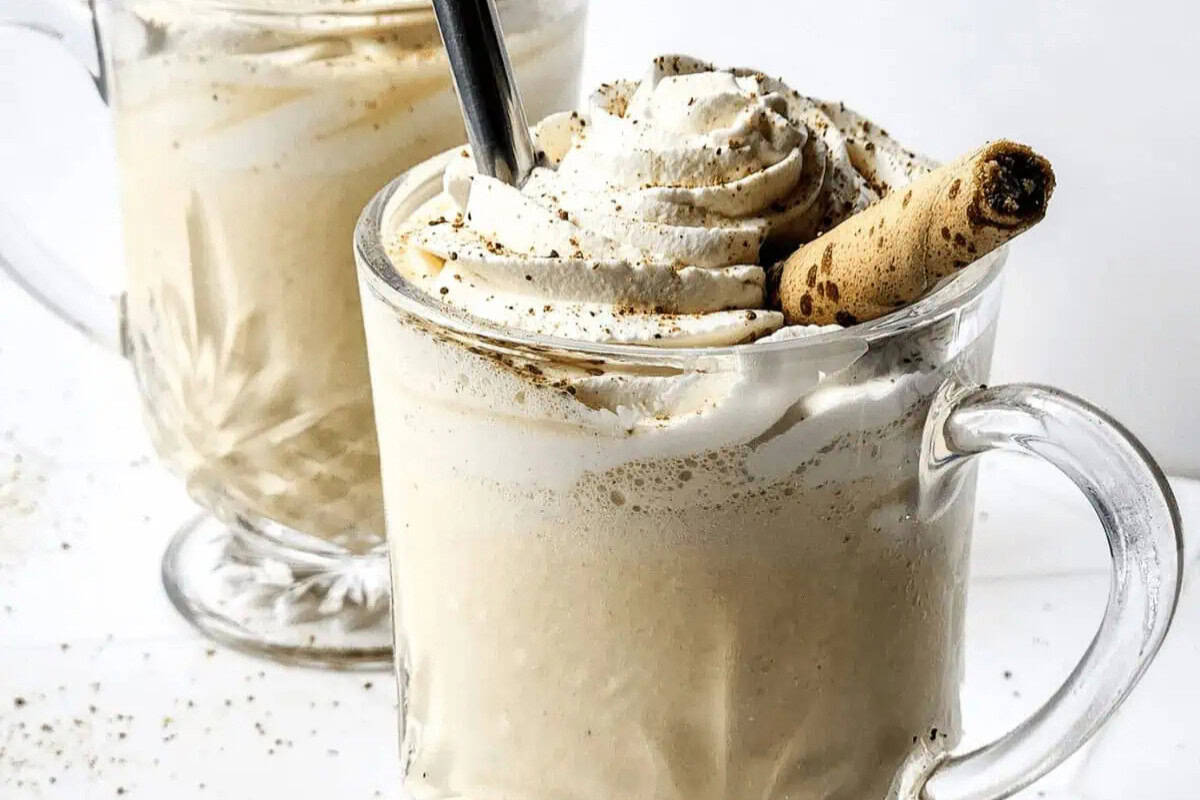 Two glass mugs filled with a creamy beverage topped with whipped cream, a rolled wafer cookie, and a straw on a white surface—an irresistible dessert perfect for summer.
