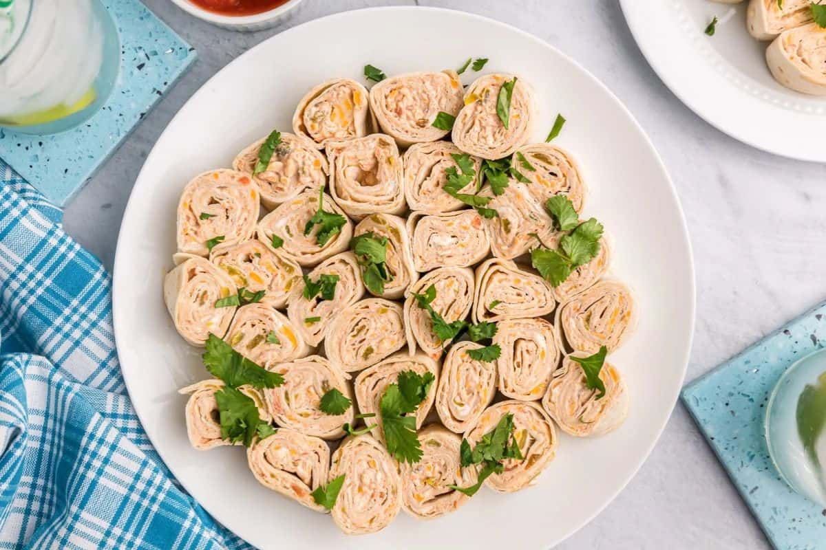 A white plate with rolled tortilla pinwheels filled with a creamy spread, garnished with fresh cilantro, on a marble surface near a blue checked napkin and drinks—perfect for those seeking cold appetizer ideas.