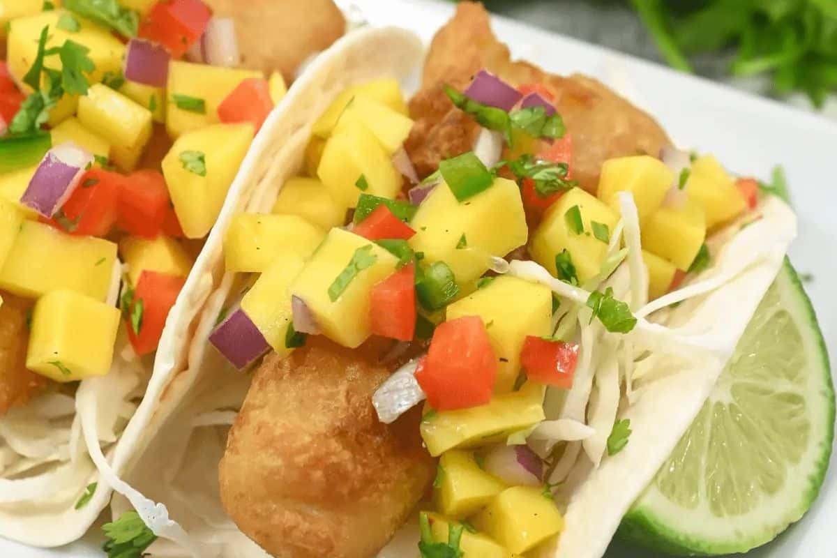 A plate of fish tacos, topped with a mango salse and coleslaw, served with a lime wedge on the side.