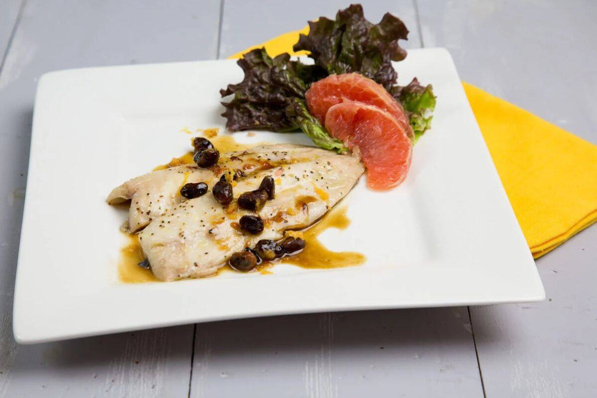 A white fish fillet with sauce and dark capers on a square plate, garnished with lettuce and grapefruit—a perfect pick for grapefruit recipes—next to a yellow napkin on a light wooden surface.