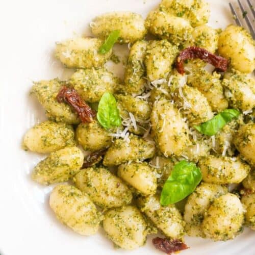 A plate of gnocchi topped with bold pesto sauce, grated cheese, sun-dried tomatoes, and fresh basil leaves, with a fork on the side.