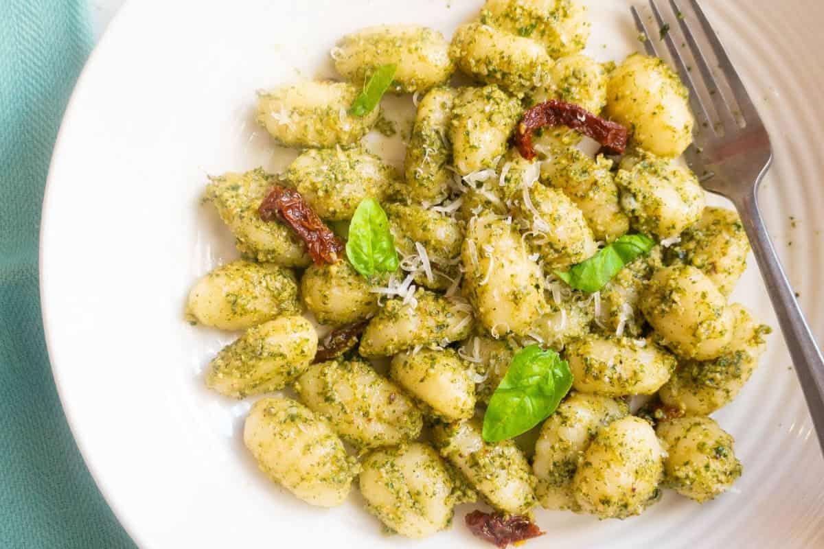 A plate of gnocchi topped with bold pesto sauce, grated cheese, sun-dried tomatoes, and fresh basil leaves, with a fork on the side.
