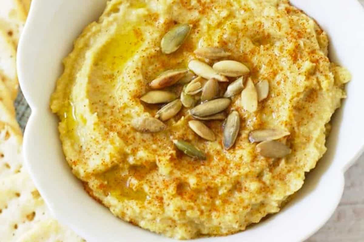 A bowl of classic hummus topped with olive oil, pumpkin seeds, and a sprinkle of paprika, served with pita bread—a delicious twist on traditional hummus recipes.