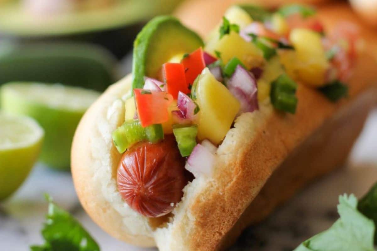 A close-up of a hot dog in a bun, topped with mango salse, avacado, white onion, and peppers.