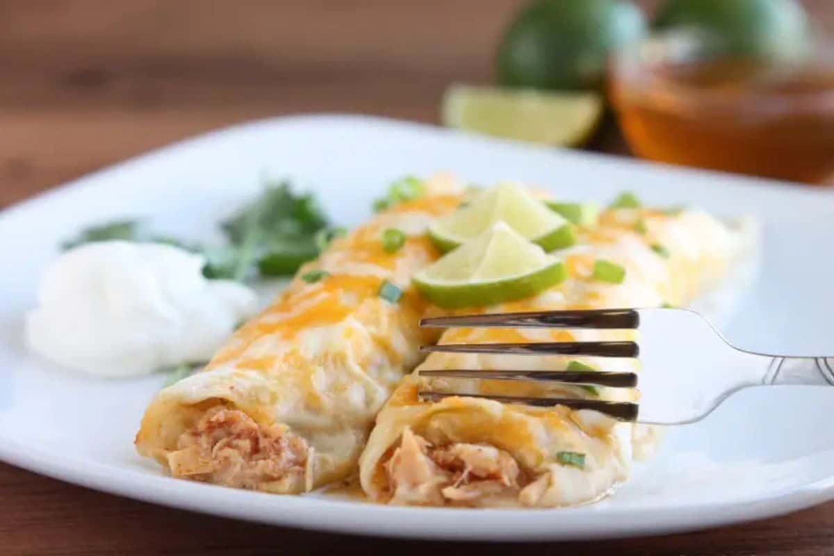 Two chicken enchiladas, made with canned chicken for easy dinner recipes, topped with melted cheese, chopped green onions, and lime wedges on a white plate, with a fork, sour cream, and cilantro on the side.