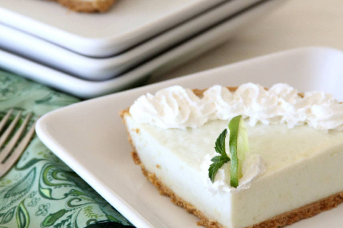 A slice of key lime pie with whipped cream and a lime garnish on a white plate, next to a green napkin and a fork—this classic treat is one of summer’s most irresistible desserts.