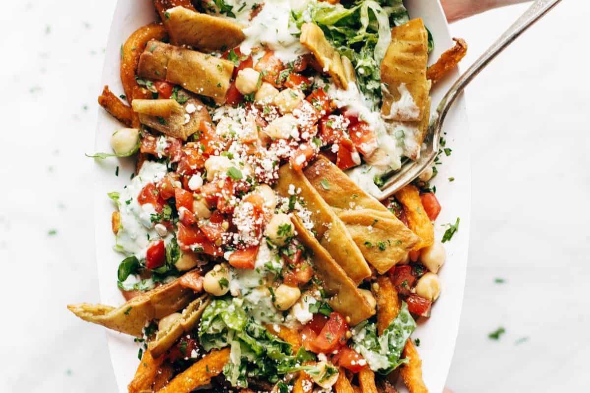 A bowl of loaded fries bursting with flavor—topped with chopped vegetables, chickpeas, crumbled cheese, pita bread slices, herbs, and a drizzle of white sauce—with a fork on the side. It's one of those French fry recipes that's crazy good!.