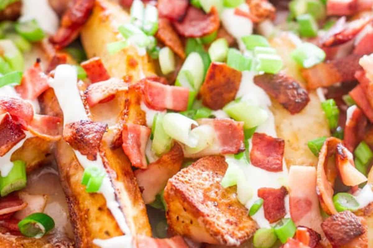 Close-up of loaded potato wedges topped with melted cheese, chopped bacon, sliced green onions, and a drizzle of white sauce—this is crazy good flavor at its best for fans of French fry recipes.