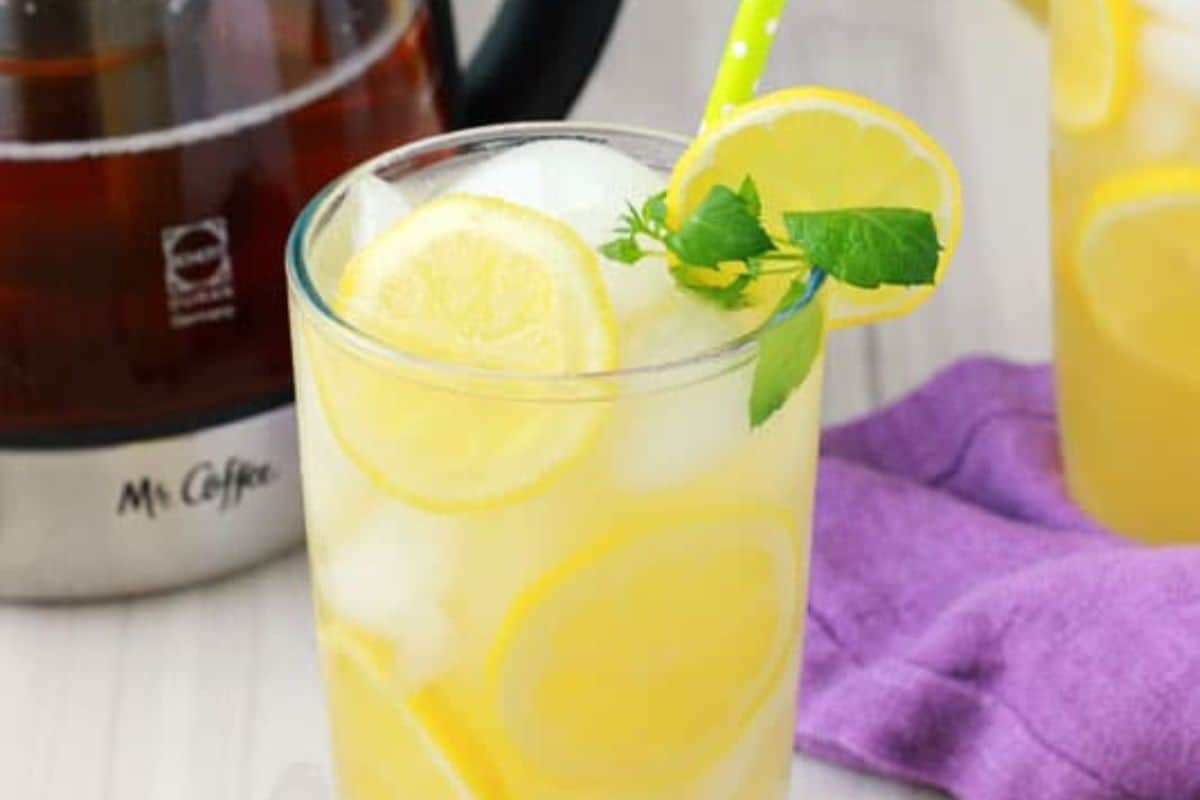 A close-up of a glass of mango green tea lemonade, garnished with mint and lemon slices, served with a yellow straw.