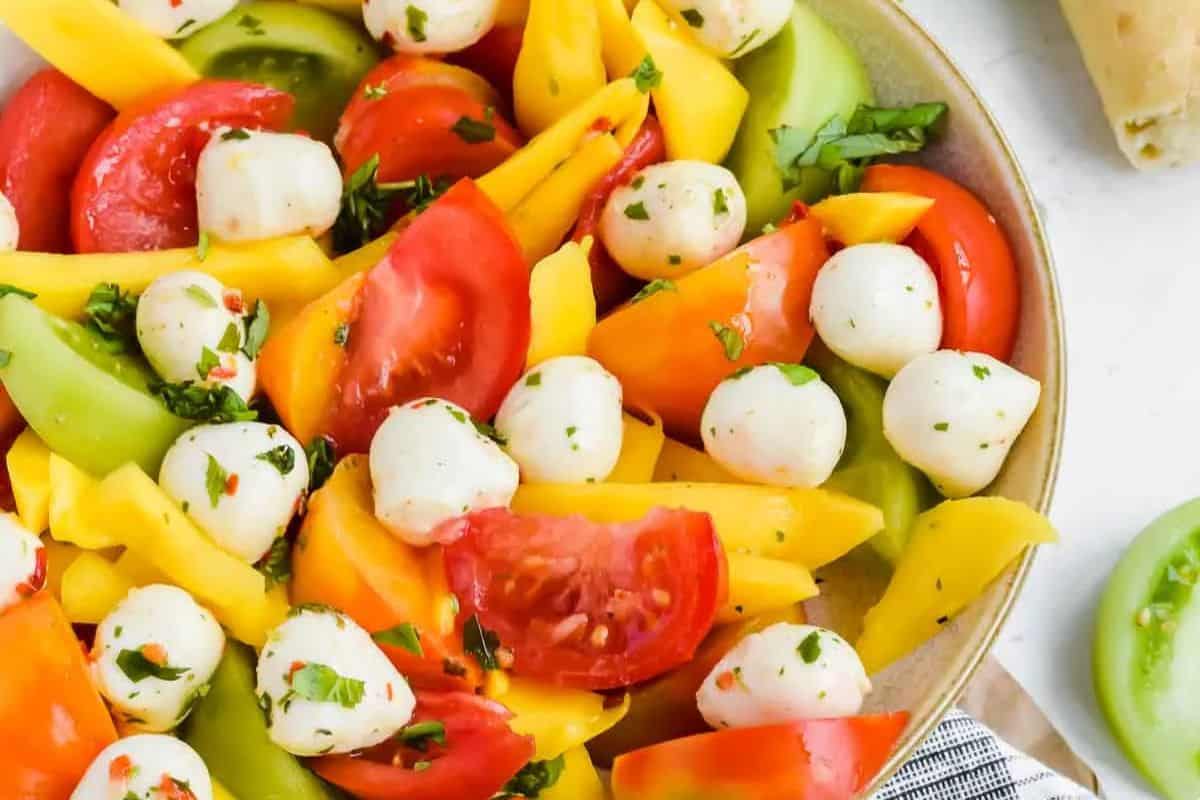 A close-up of a bowl of mango tomato salad, with mozzarella cheese, green and red tomatoes, and mangos.