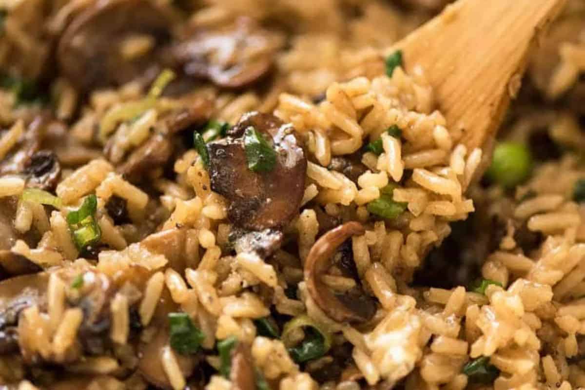 Close-up of cooked rice mixed with sliced mushrooms and chopped green onions, being stirred with a wooden spoon—a must try for fans of mushroom dishes looking to go beyond the basics.