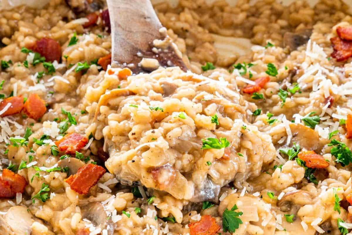 A close-up of a creamy risotto, one of the must try mushroom dishes, parsley, and grated cheese, being stirred with a wooden spoon.