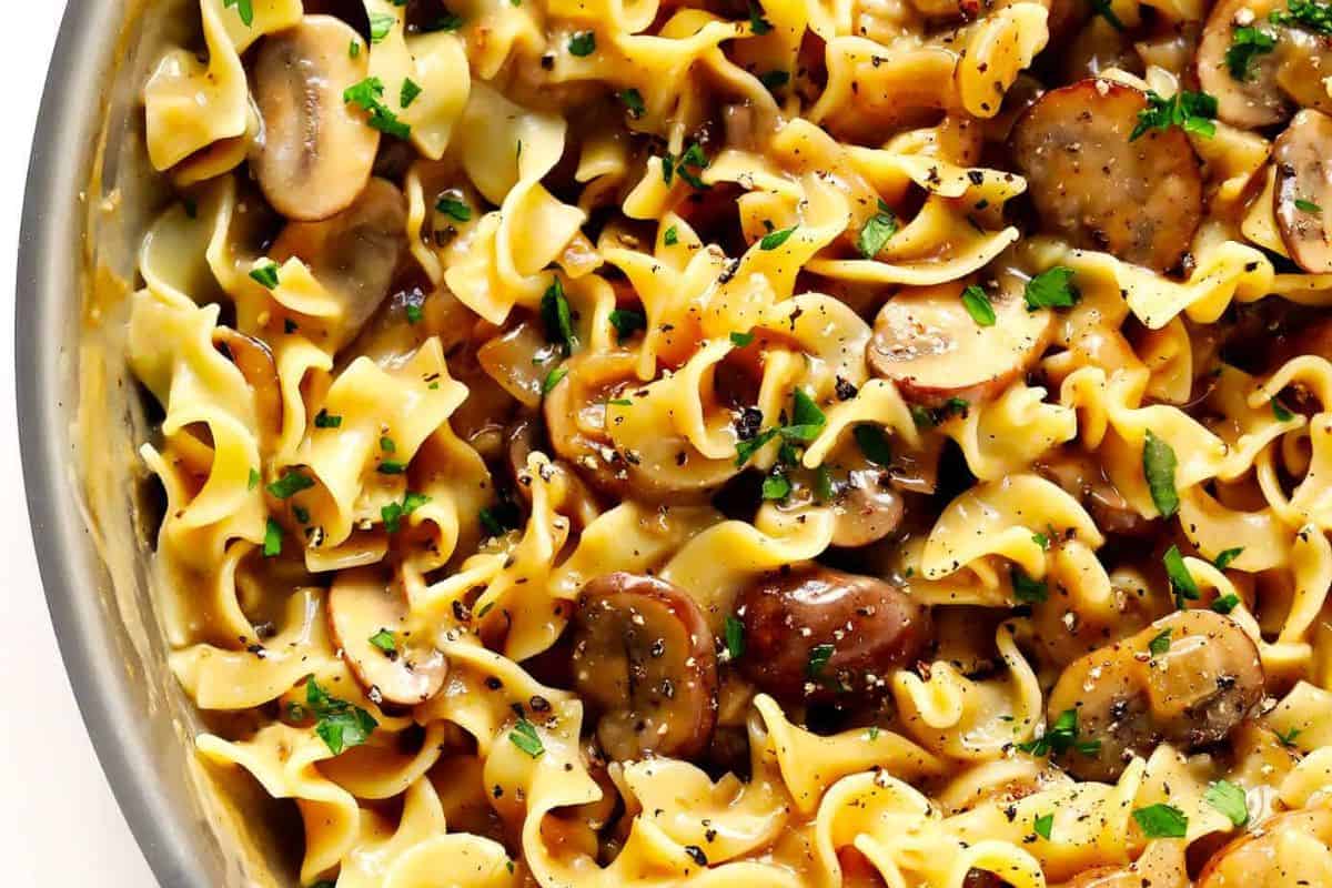 A close-up of cooked egg noodles mixed with sliced mushrooms, garnished with chopped parsley and ground black pepper—a mushroom dish that's a must try for those looking to go beyond the basics.